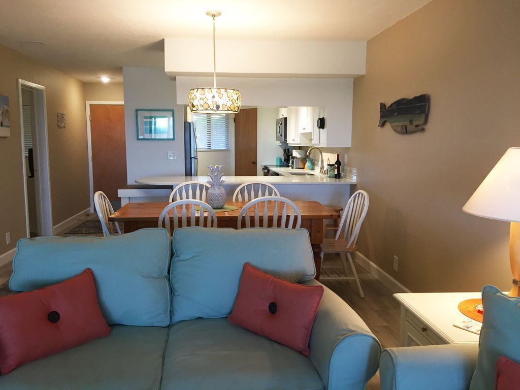 Una sala moderna con un sofá azul claro, cojines rojo y negro, y un comedor con una mesa de madera y sillas blancas.