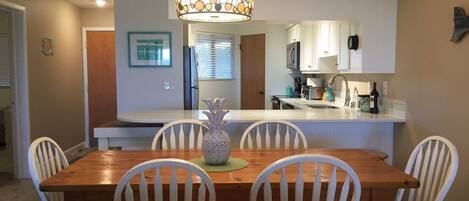 Un comedor con una mesa de madera y sillas blancas, un jarrón decorativo, y una cocina al fondo.