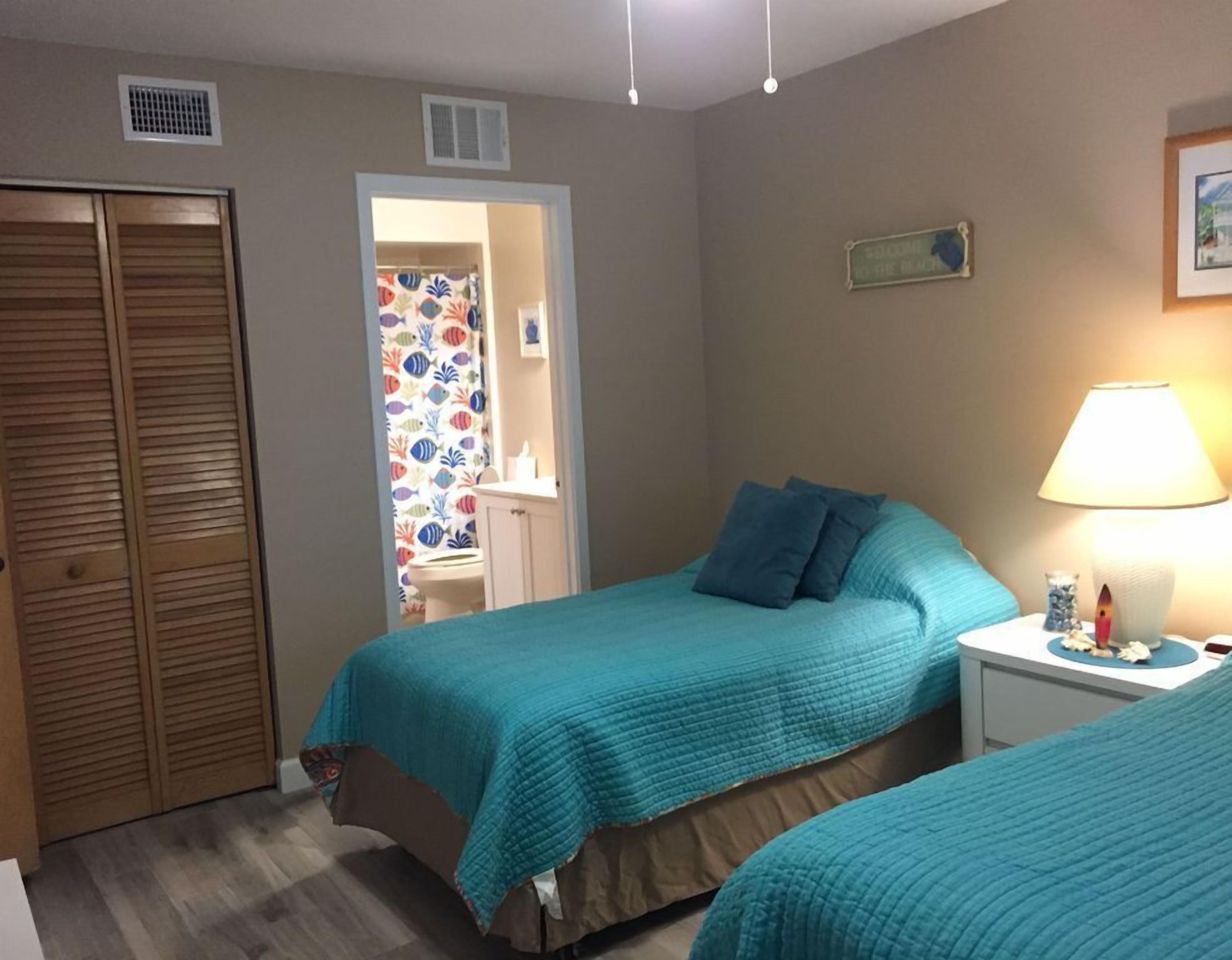 A bedroom with two beds, a nightstand, a lamp, a wardrobe, and a bathroom visible through an open door.
