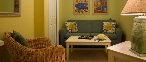 A living room with a green sofa, a wicker chair, a coffee table, and two paintings on the wall.