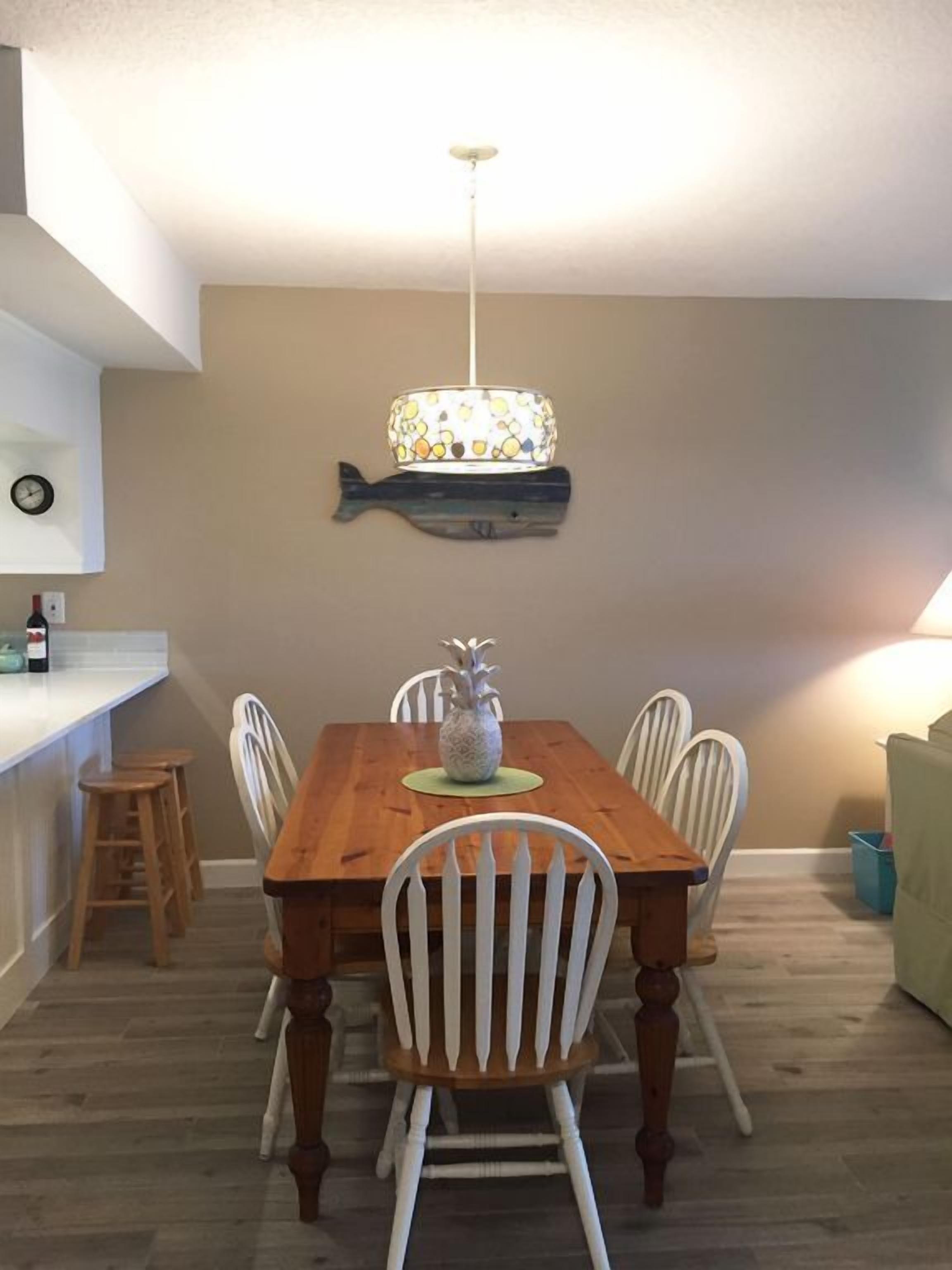 Un coin repas avec une table en bois, des chaises blanches, un luminaire décoratif en forme de poisson et une horloge au mur.
