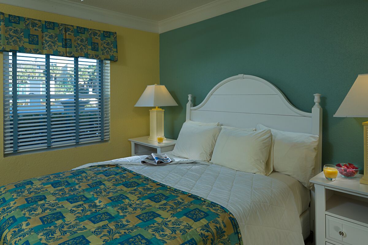 A neatly made bed with a patterned bedspread, two bedside lamps, a nightstand with a glass of orange juice, and a window with blinds.
