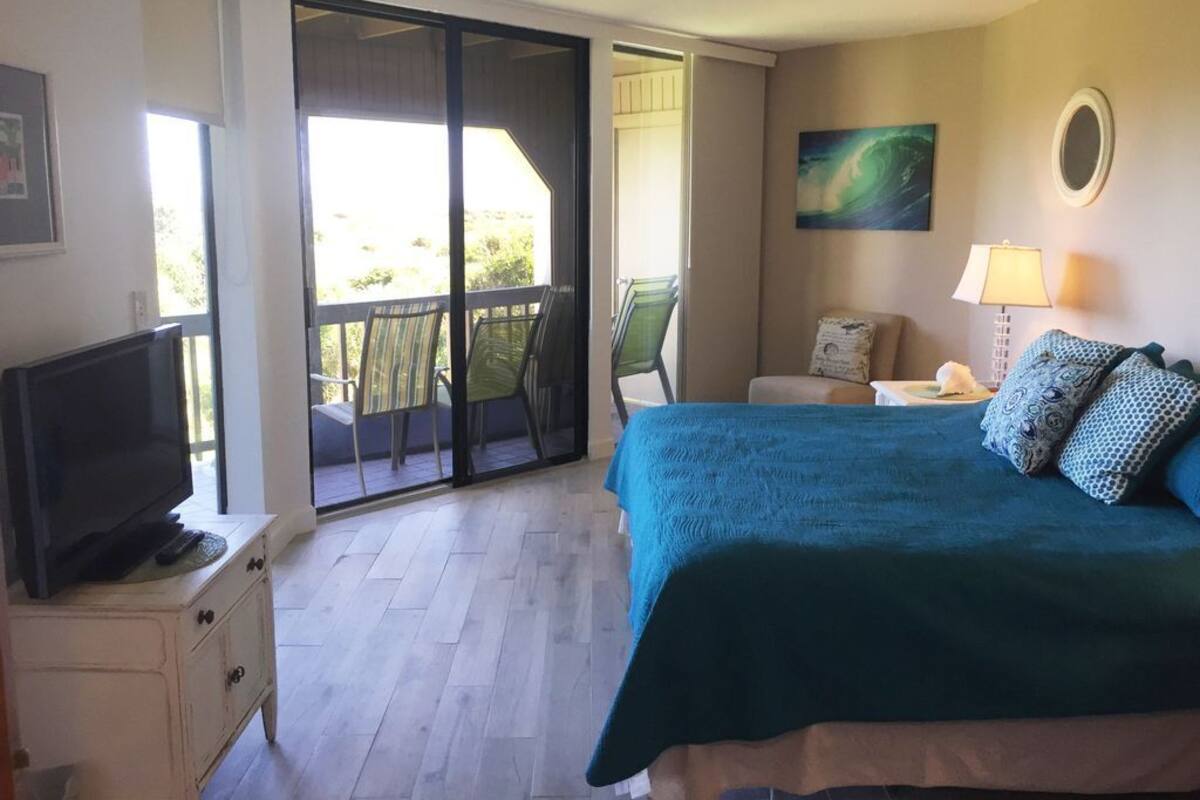 A bedroom with a bed, a television on a dresser, a balcony with chairs, and a painting on the wall.