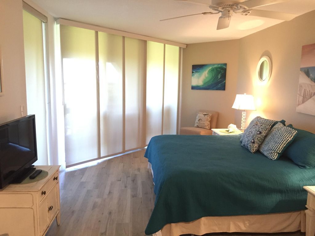 Un dormitorio con una cama, un televisor sobre un aparador, un ventilador de techo y una ventana con persianas.