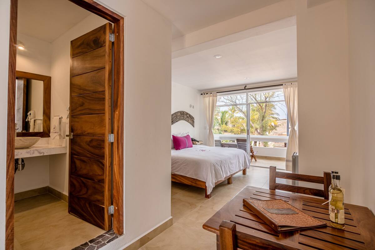 Un dormitorio con puerta de madera, cama con ropa blanca, ventana con cortinas y una mesa con una botella.