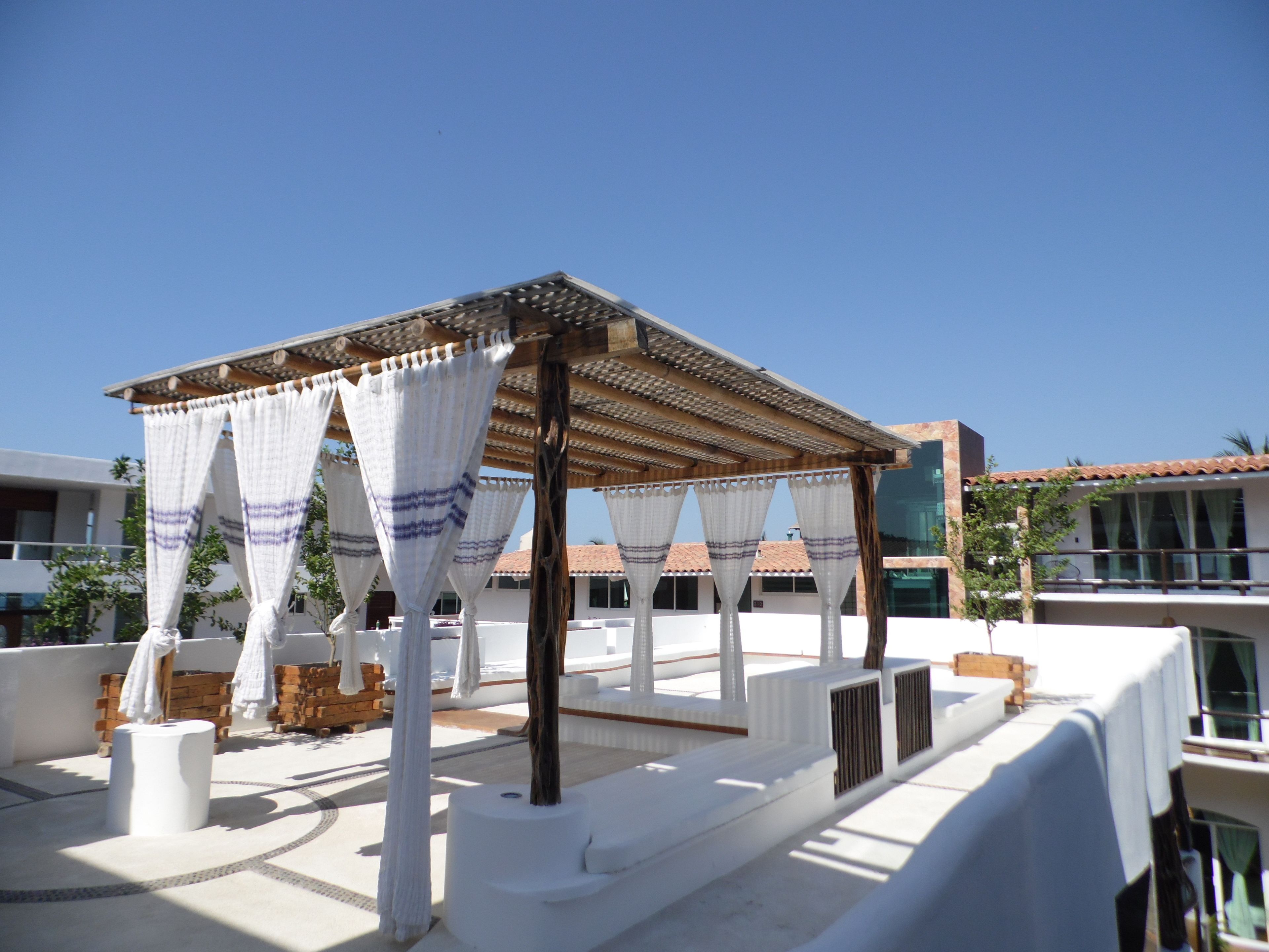 Una terraza en la azotea con una pérgola, cortinas blancas y mobiliario exterior.