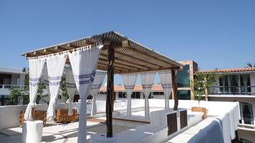Una terraza en la azotea con una pérgola, cortinas blancas y mobiliario exterior.