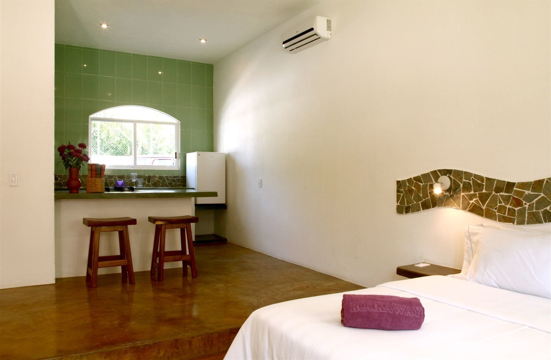 A hotel room with a bed, a nightstand, a stone headboard, a window with a view, two wooden stools, and a kitchen area with a refrigerator and a window.