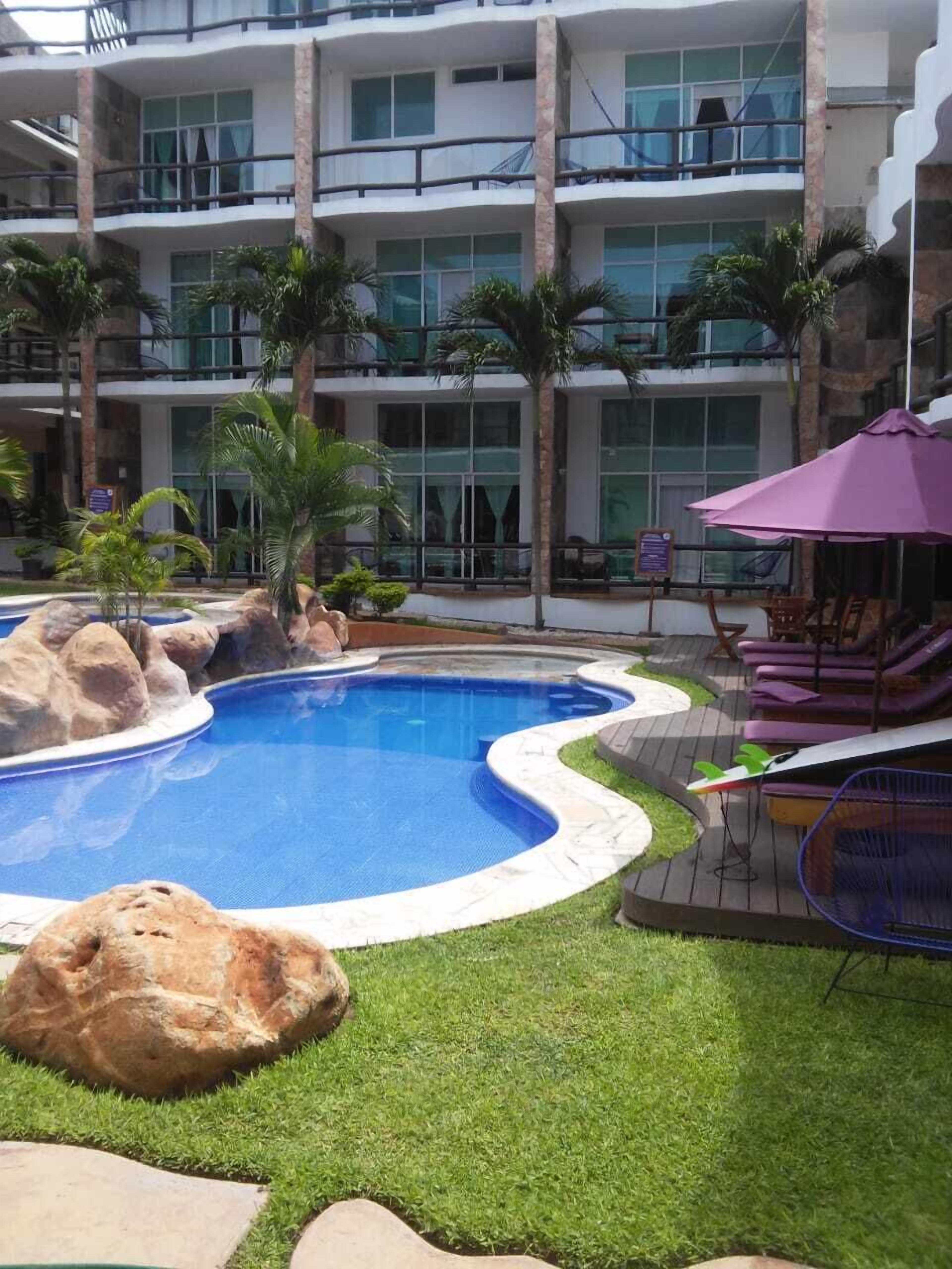 A swimming pool with a stone border, surrounded by grass and palm trees, with a building in the background.