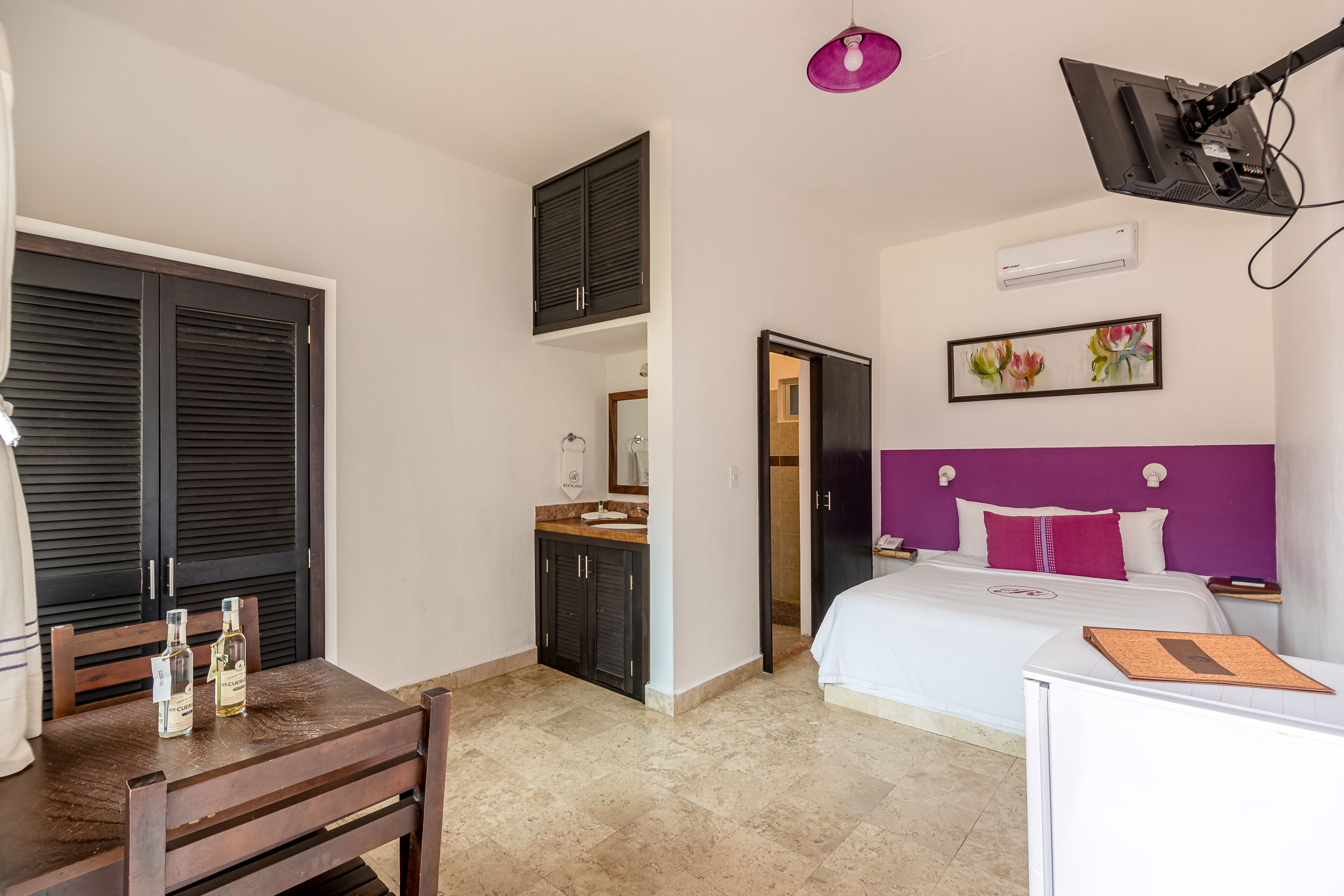 Ein Hotelzimmer mit einem Bett, einem Schreibtisch, einem Sessel, einem Tisch mit zwei Flaschen, einem Spiegel und einem Fenster mit Fensterläden.