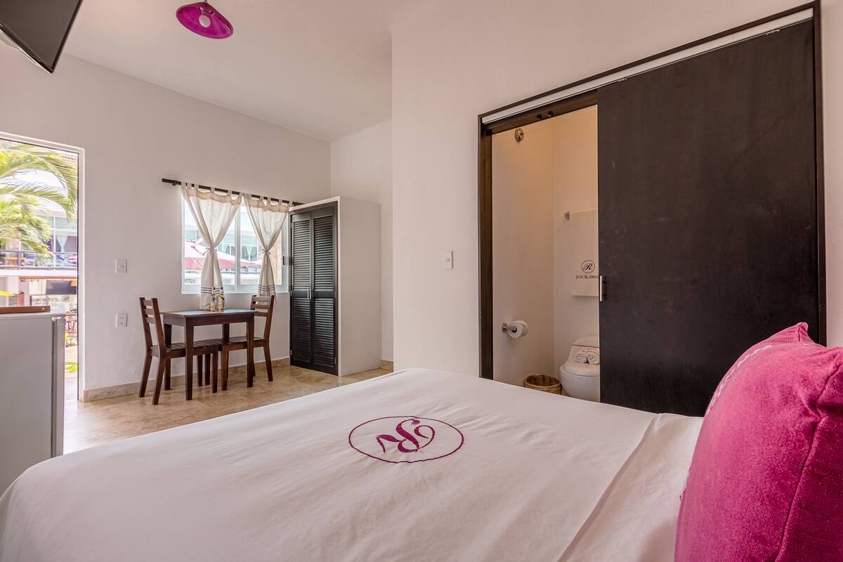 Una habitación de hotel con cama, una mesa de comedor, sillas, una ventana con cortinas y una puerta.