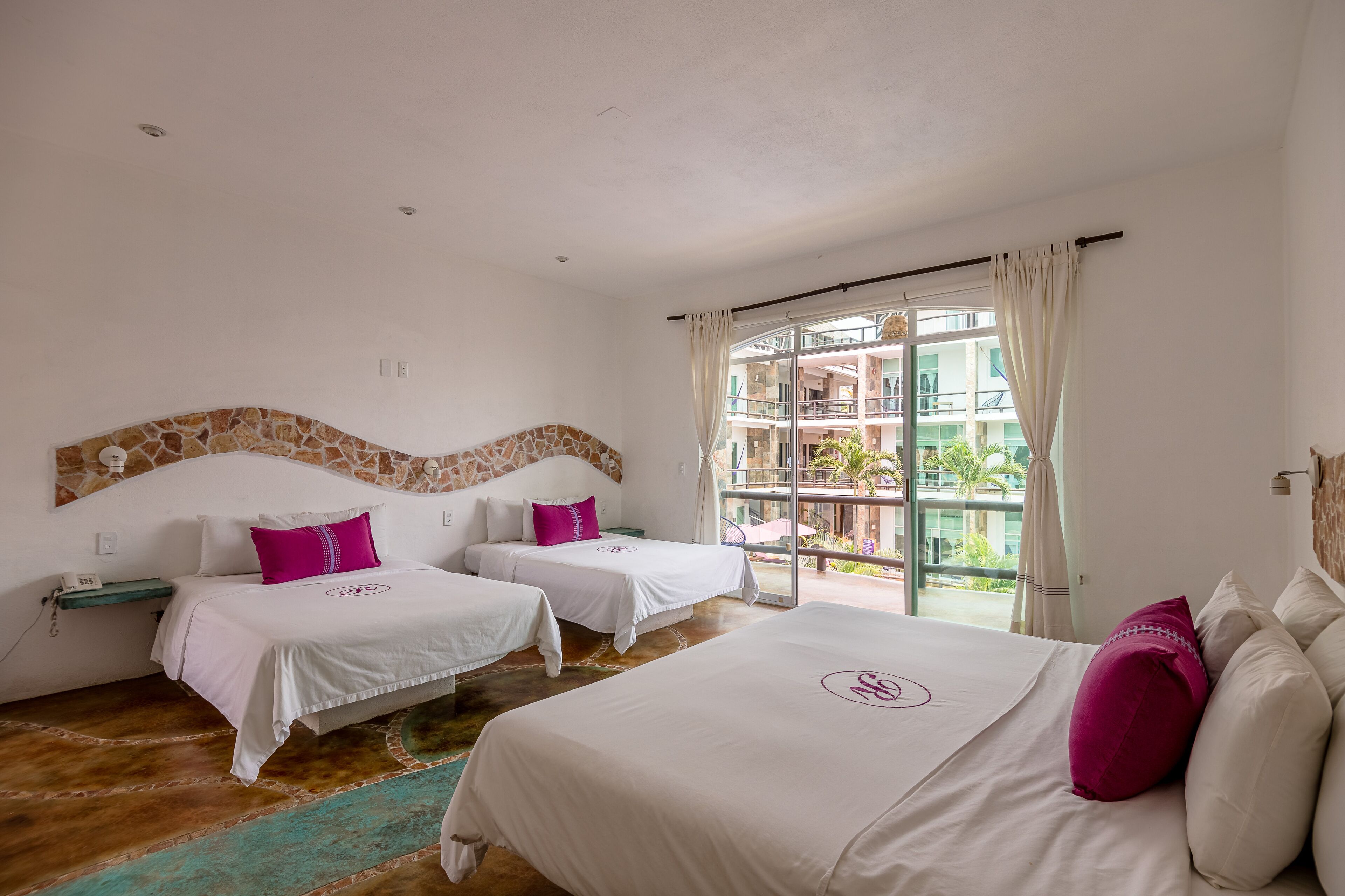 A hotel room with two beds, a large window with a view of a building and palm trees, and a stone accent wall.