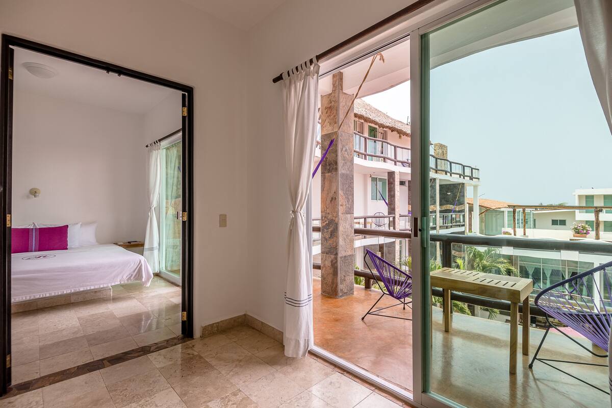 Una habitación de hotel con cama, balcón con mesa y sillas, y vistas de los edificios circundantes.