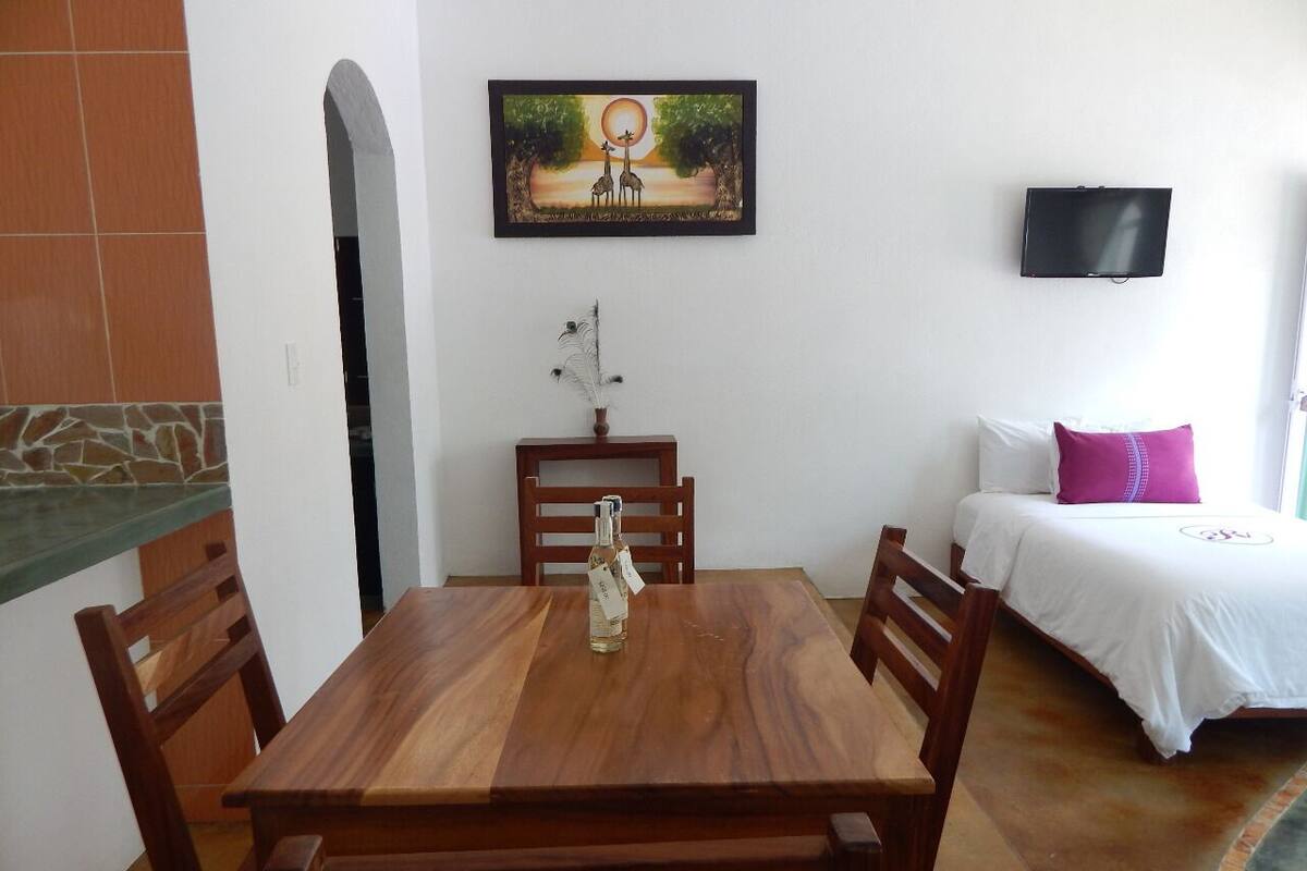 Un comedor con una mesa y sillas de madera, un cuadro sobre la mesa, y una cama con un televisor al fondo.