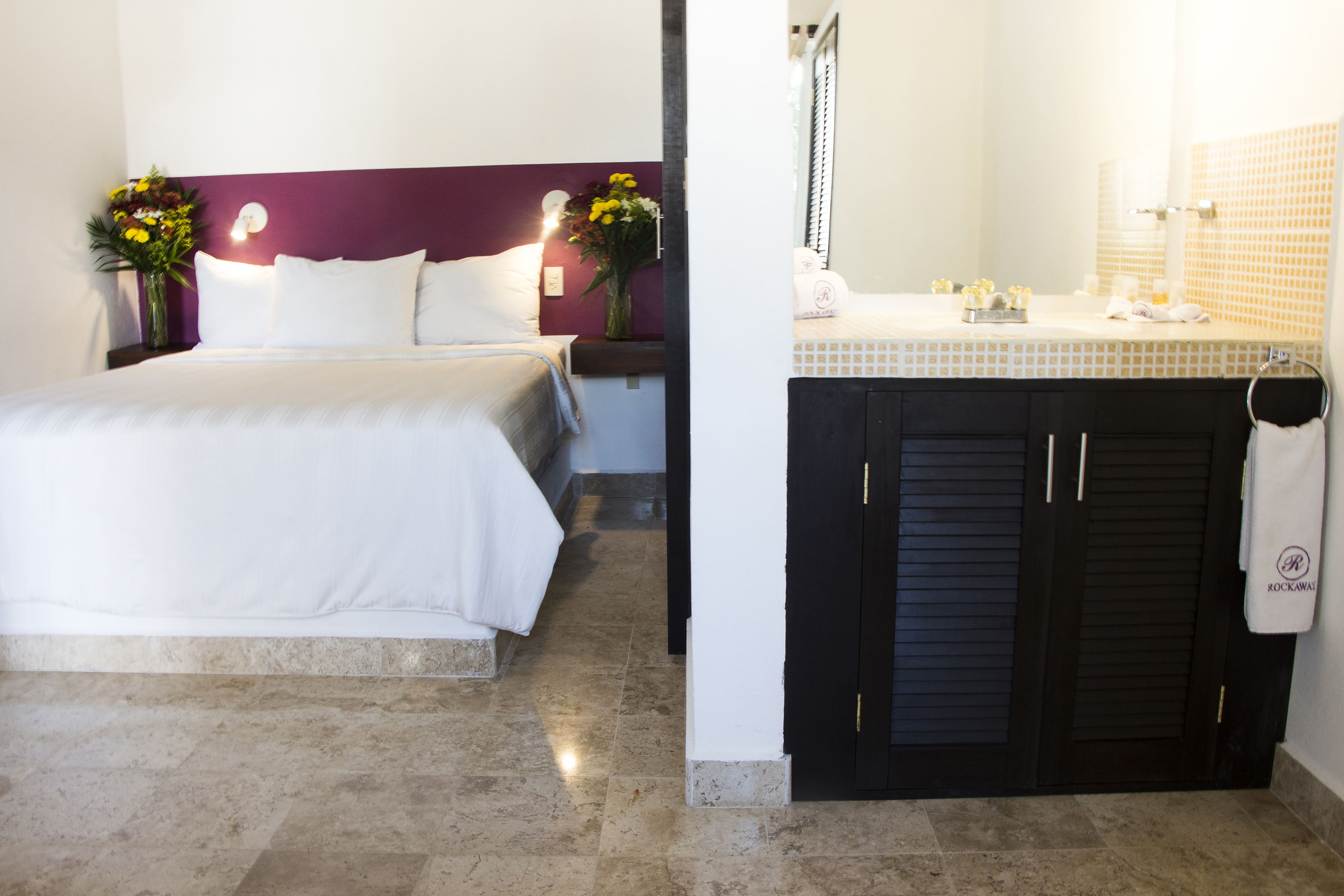 A hotel room with a bed, a nightstand with a vase of flowers, a bathroom with a sink and mirror, and a towel rack.