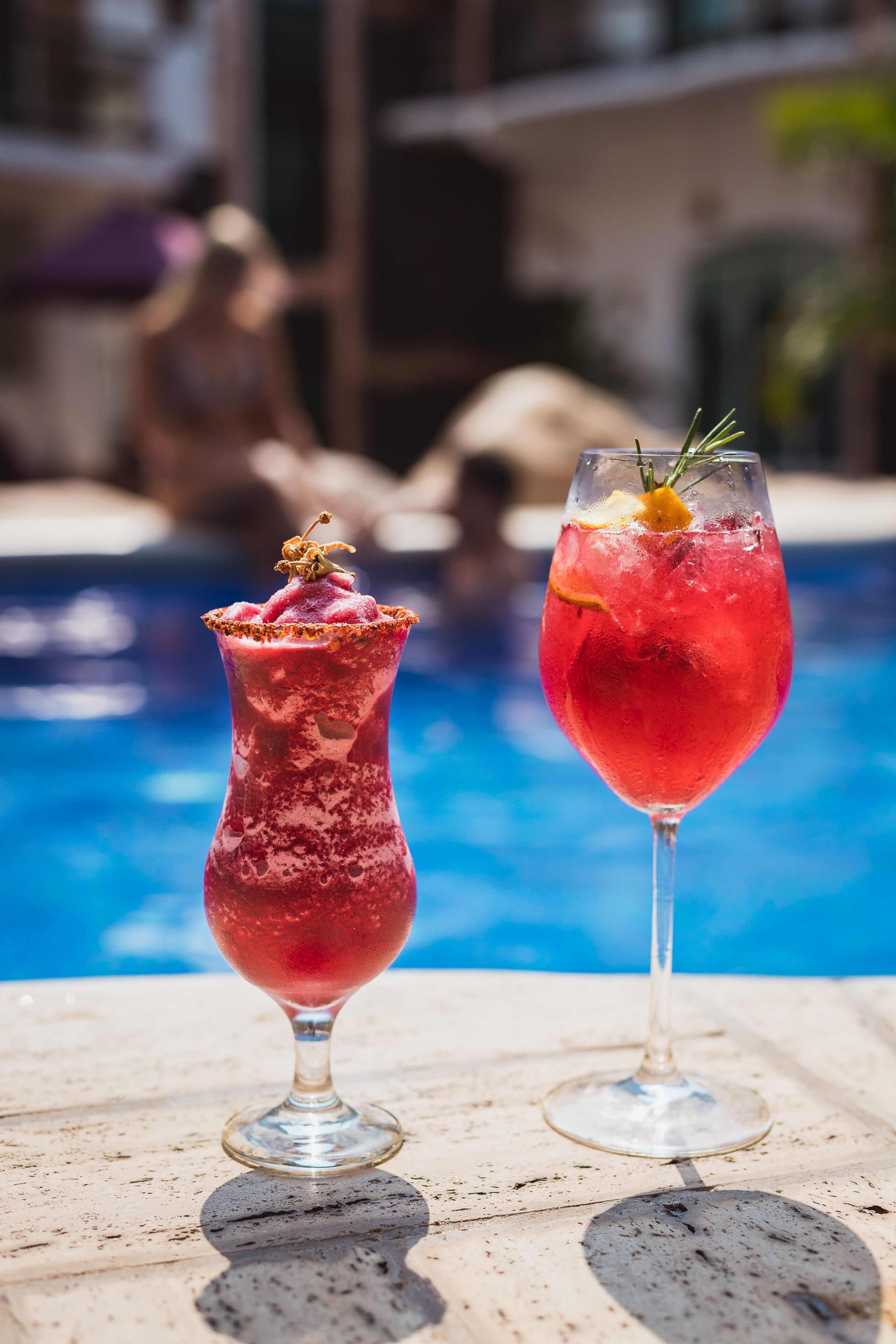 Two cocktails on a table by a poolside with a blurred background of people and buildings.