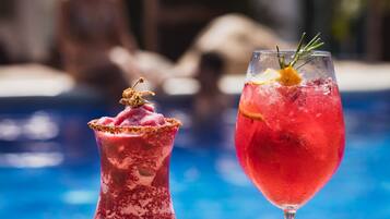 Two cocktails on a table by a poolside with a blurred background of people and buildings.