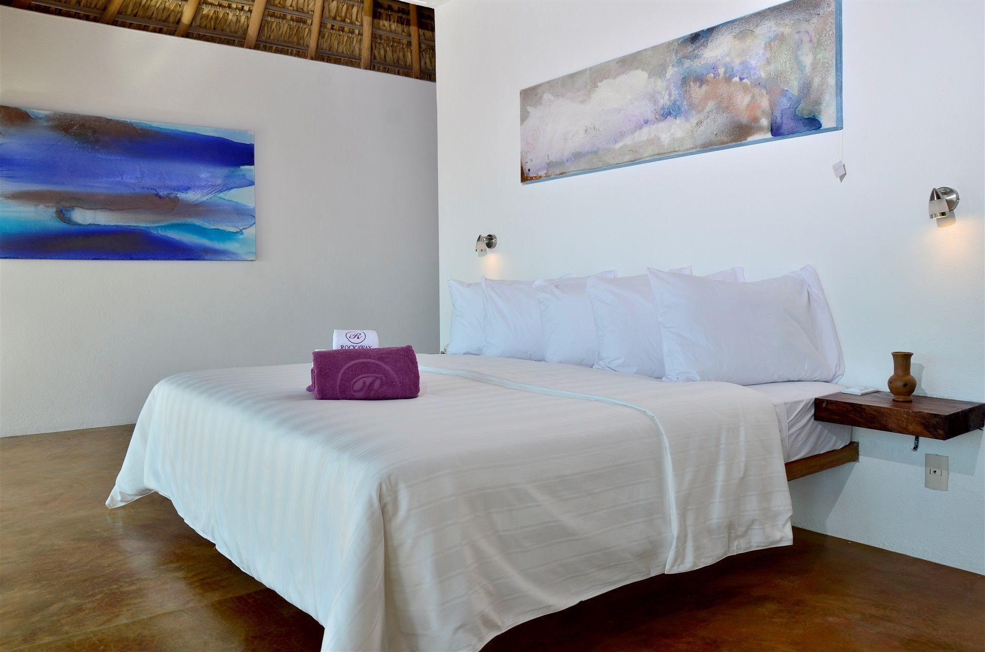A hotel room with a bed, a painting on the wall, and a small table with a vase.