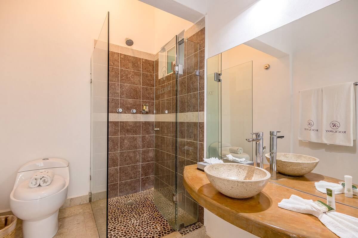 Un baño con ducha de vidrio, inodoro blanco y tocador de madera con dos lavamanos grandes.