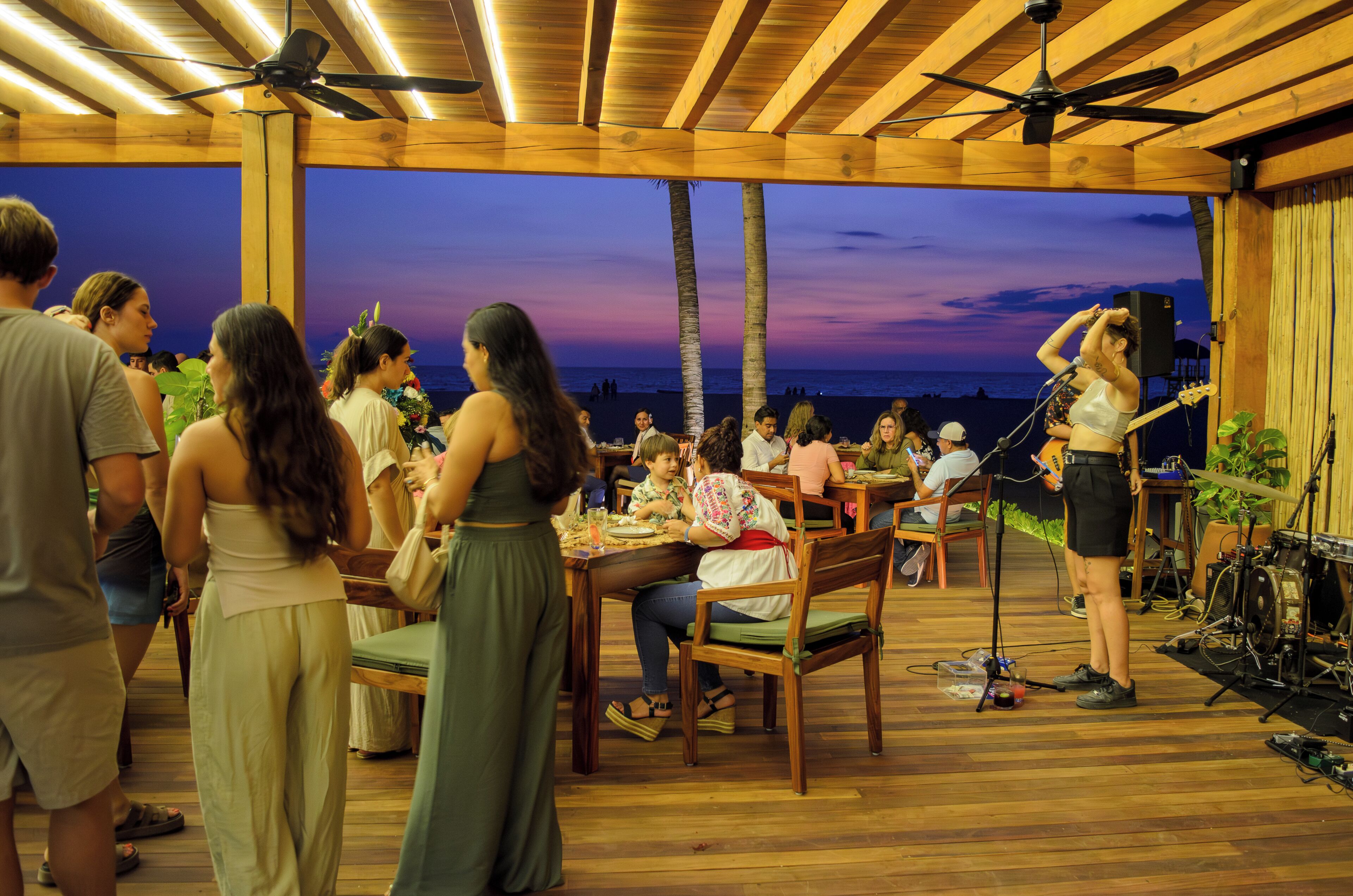 An outdoor dining area with wooden tables and chairs, a stage with musical equipment, and a view of the ocean at dusk.
