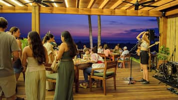 An outdoor dining area with wooden tables and chairs, a stage with musical equipment, and a view of the ocean at dusk.
