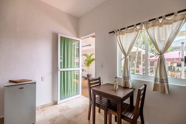 Habitación compacta con una mesa de comedor, sillas, un refrigerador pequeño y una puerta corrediza de vidrio que da a un área exterior con vistas a un edificio y palmeras.