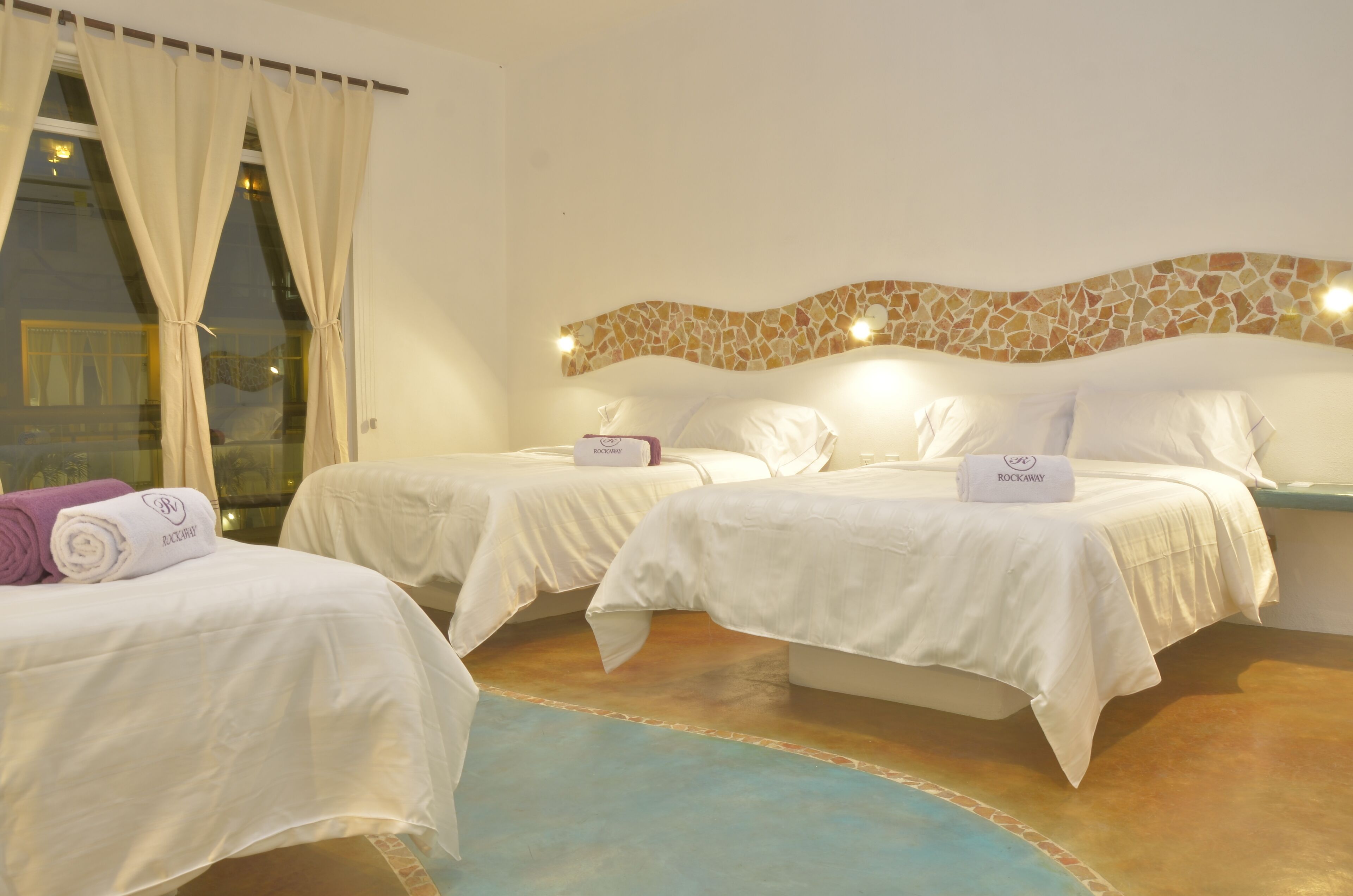 A hotel room with two beds, a stone mosaic headboard, and a view of the city through large windows.