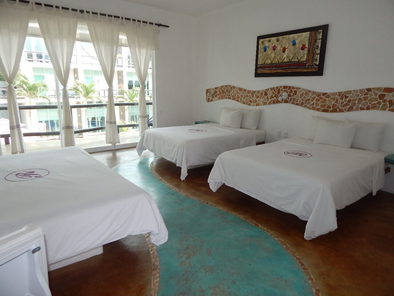 A hotel room with two beds, a large window with curtains, a mosaic headboard, and a painting on the wall.