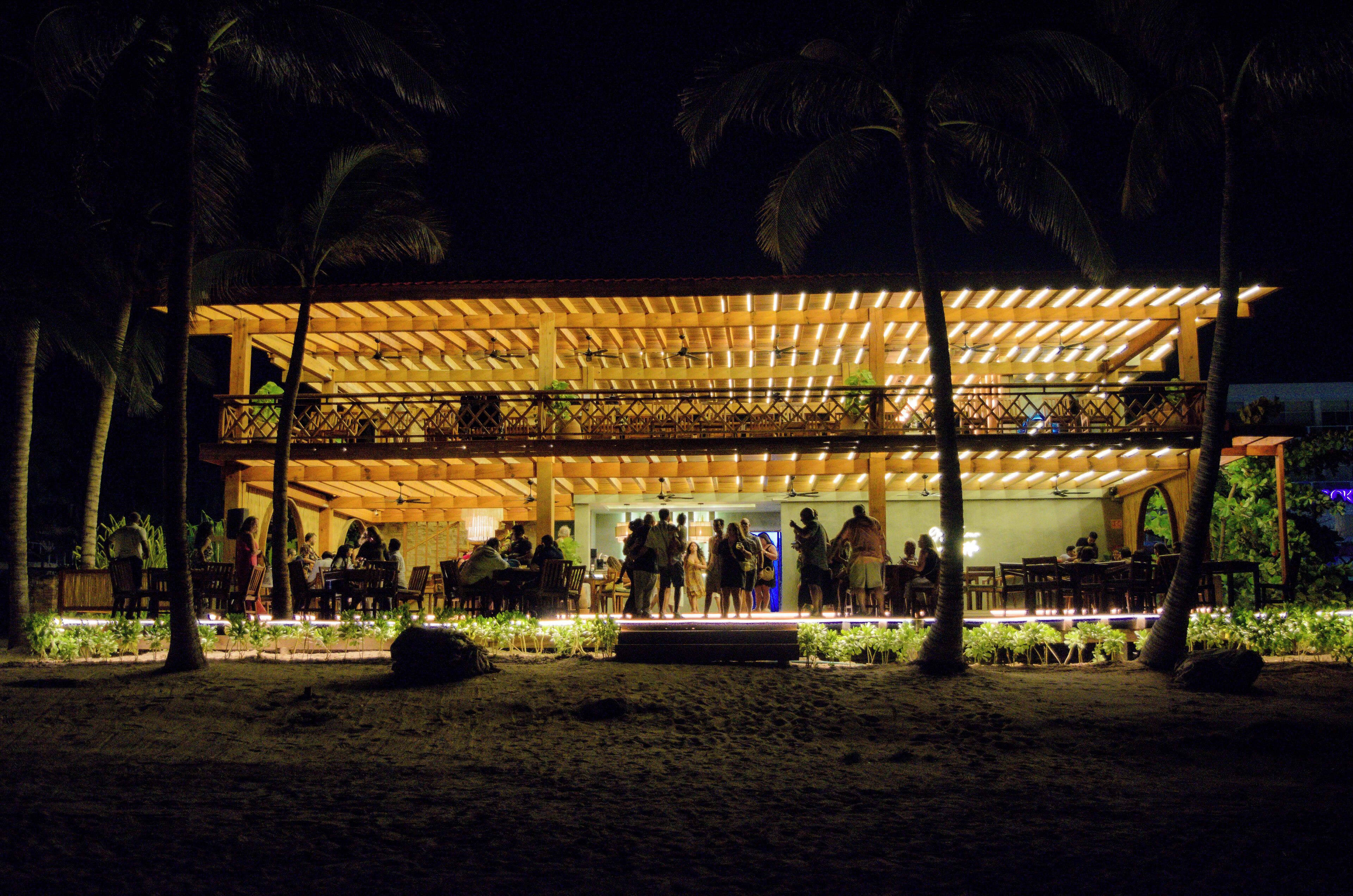 A beachside restaurant with an open-air deck, palm trees, and a sandy ground.