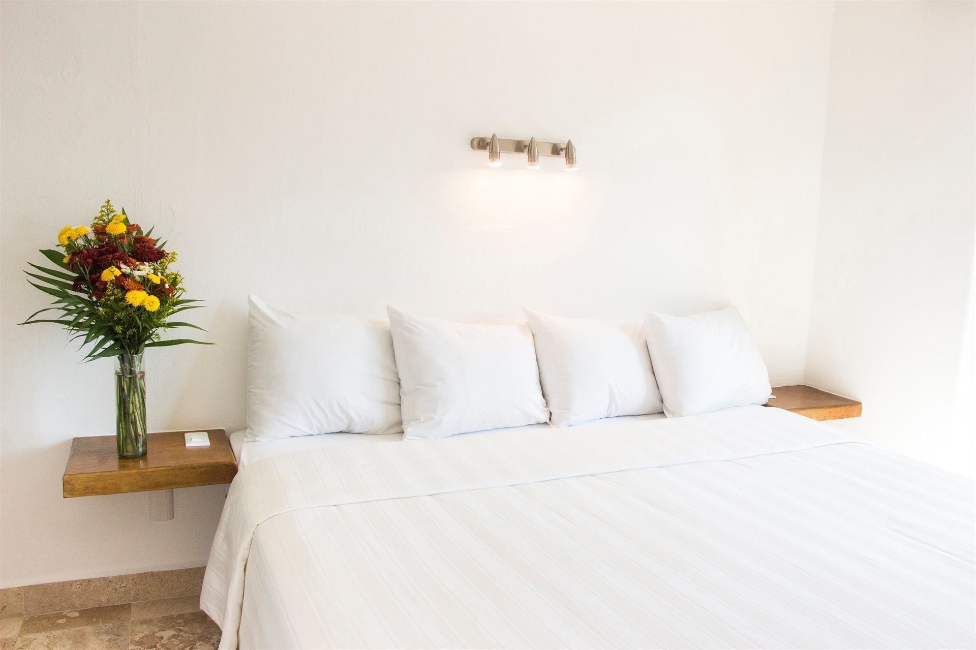 Uma cama bem arrumada com lençóis brancos, um criado-mudo de madeira com um pequeno vaso de flores e um aparelho de luz fixado na parede.