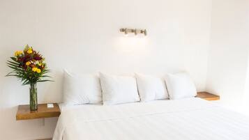 Uma cama bem arrumada com lençóis brancos, um criado-mudo de madeira com um pequeno vaso de flores e um aparelho de luz fixado na parede.