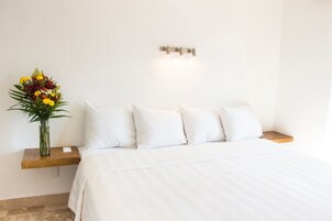Uma cama bem arrumada com lençóis brancos, um criado-mudo de madeira com um pequeno vaso de flores e um aparelho de luz fixado na parede.