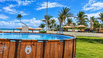 Un jacuzzi de madera rodeado por una piscina, palmeras y sombrillas de playa.