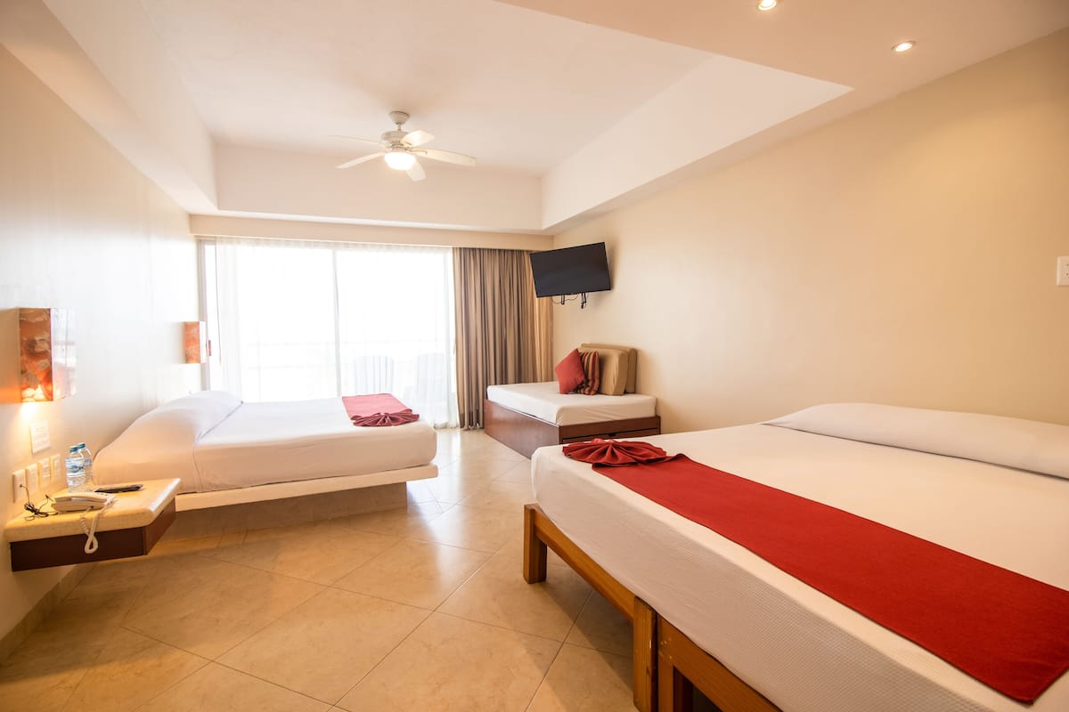 Een hotelkamer met twee bedden, een aan de muur bevestigde televisie, een plafondventilator en een groot raam met gordijnen.