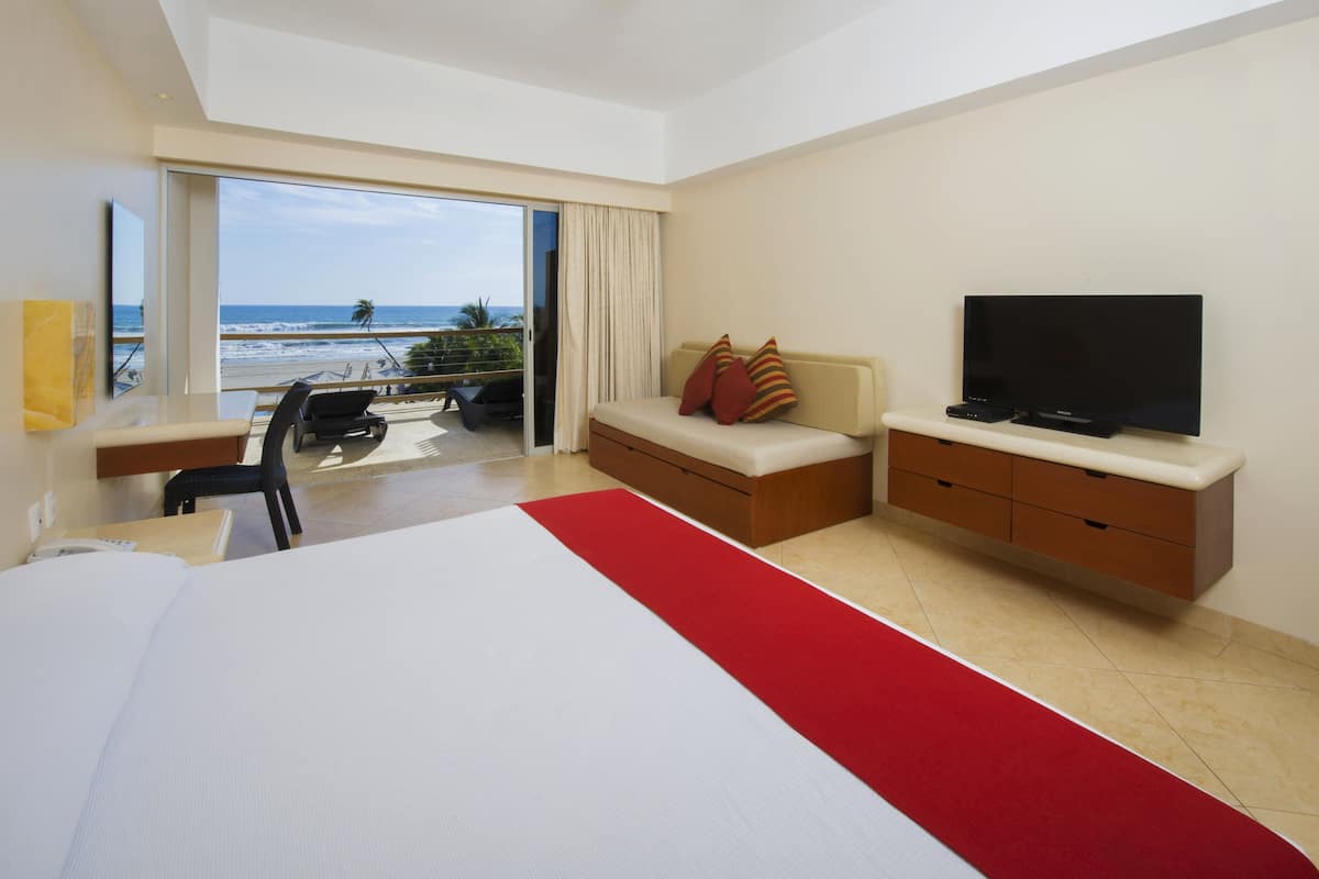 Een hotelkamer met een groot bed, een televisie, een bureau en een balkon met uitzicht op het strand.