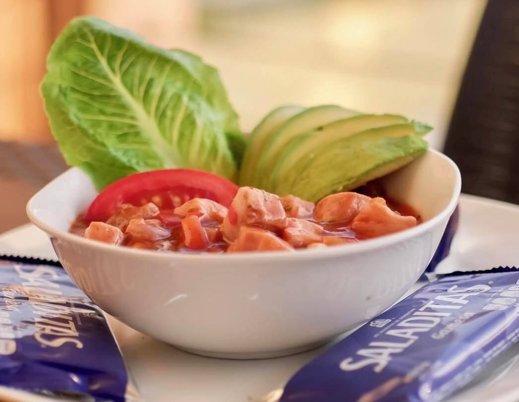 Een schaal eten met avocado, tomaat en bladgroenten, met een blauw pak erbij.