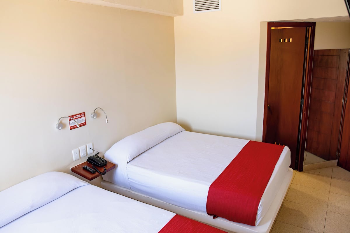 Een hotelkamer met twee bedden, een nachtkastje met een telefoon en een rode deken, en een houten deur.