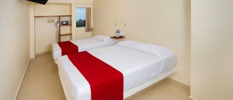 Quarto de hotel com duas camas, uma televisão fixada na parede, uma pequena escrivaninha com abajur e uma janela com cortinas.