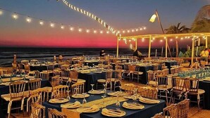 Un área de comedor al aire libre con mesas y sillas dispuestas para una comida, con vista al océano al atardecer.