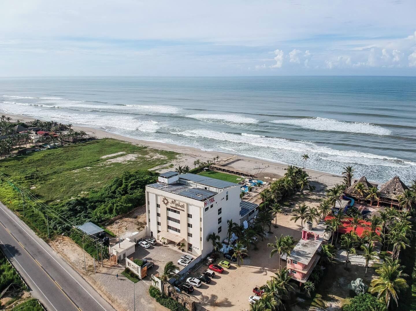 Un hôtel avec vue sur la plage, des panneaux solaires sur le toit et un parking pouvant accueillir plusieurs voitures.