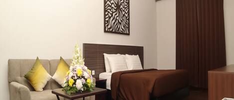 Habitación de hotel con cama, sofá, una pequeña mesa con un arreglo floral, un adorno de pared y un escritorio de madera.