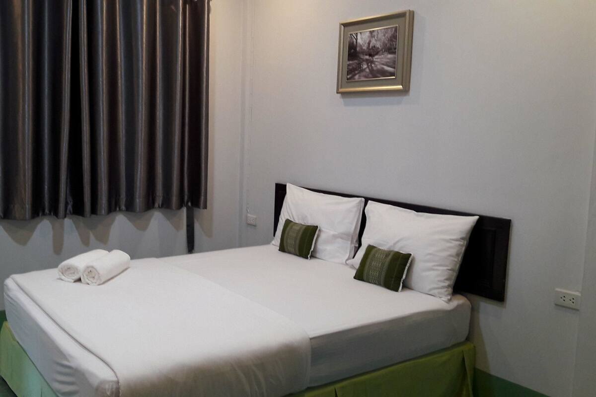 A neatly made bed with white linens and two pillows, a framed picture on the wall, and a window with dark curtains.