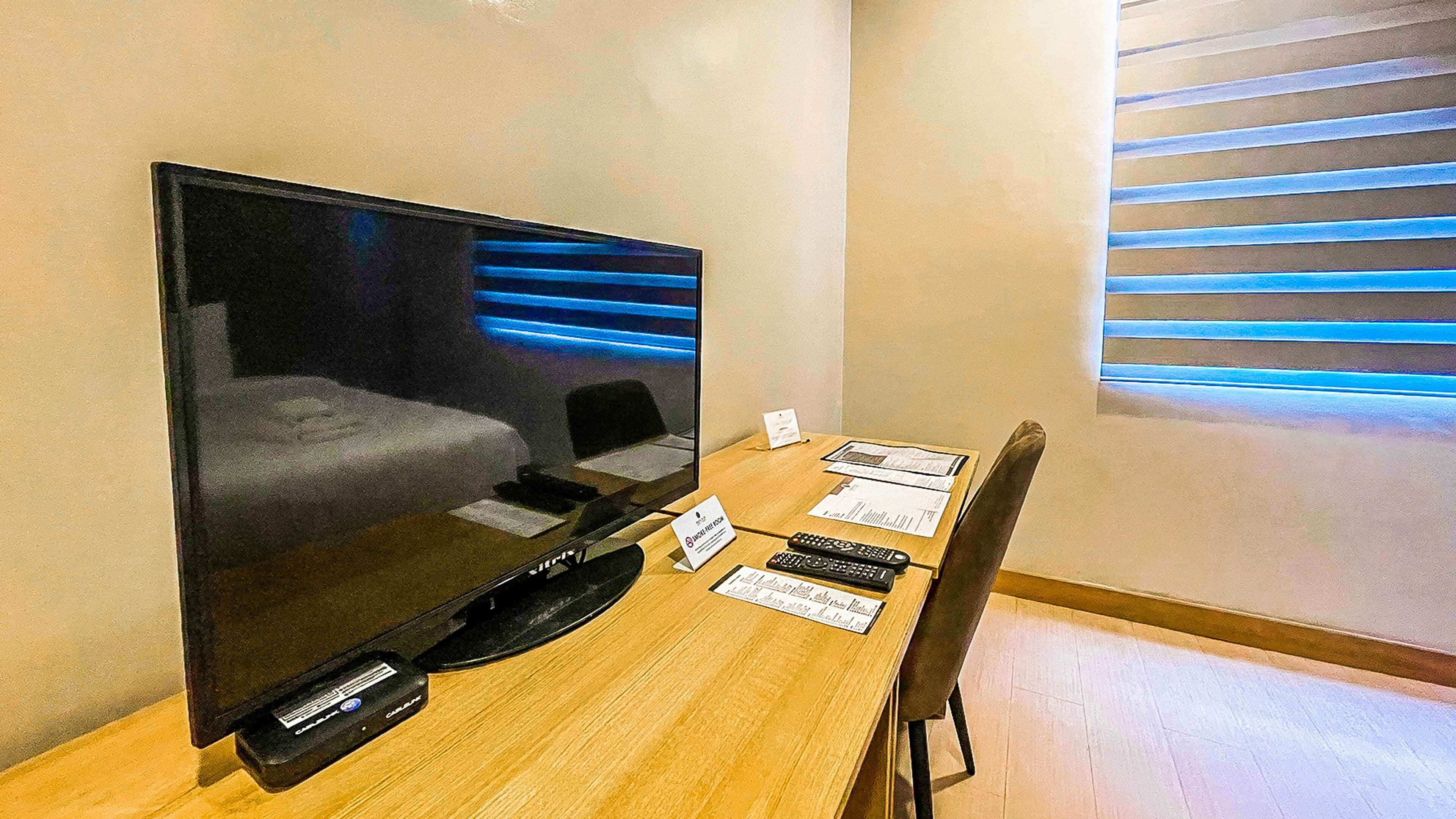A hotel room with a large flat-screen TV, a wooden table, a chair, and a window with blinds.