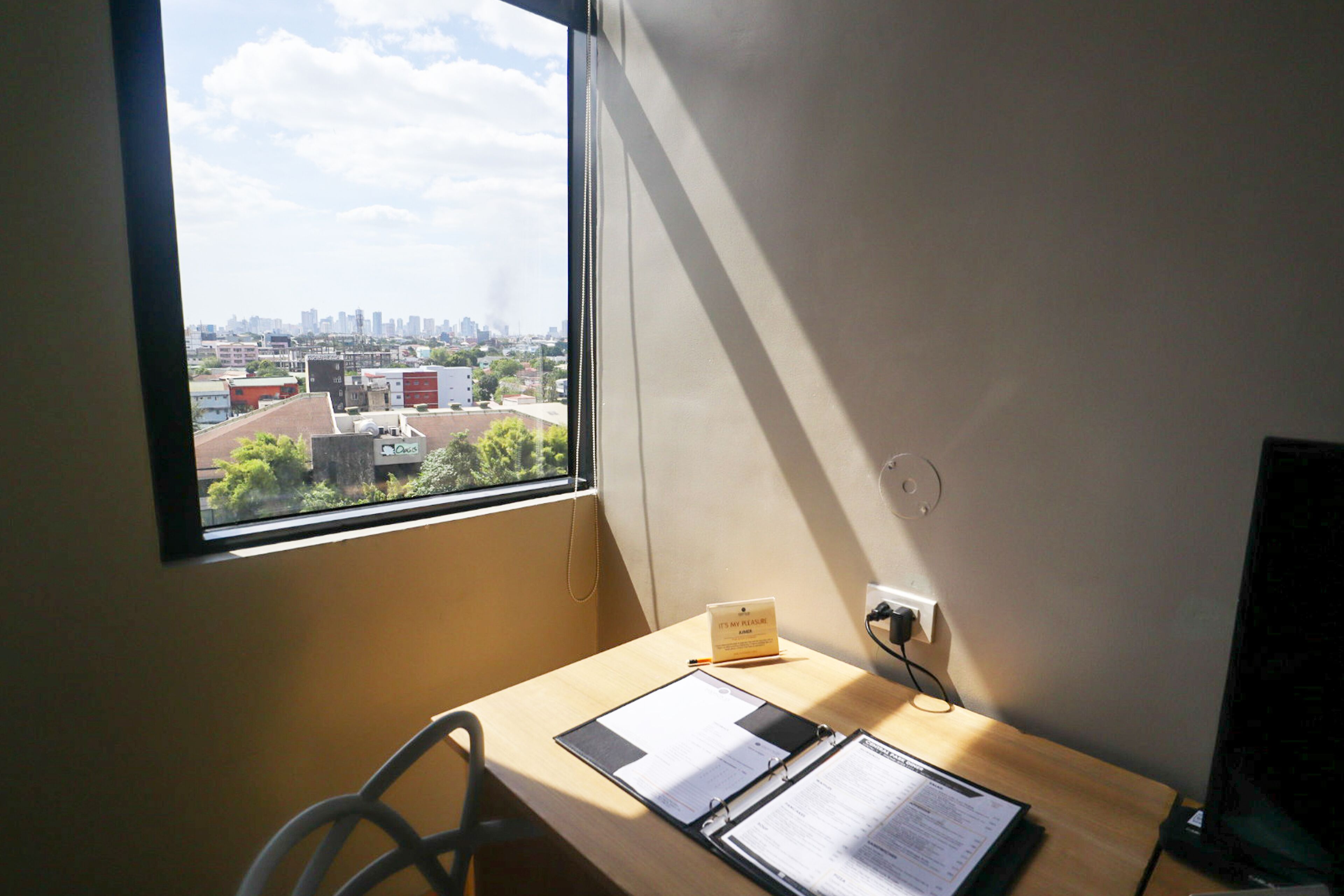 Ein Zimmer mit Schreibtisch, Stuhl, Fenster und Blick auf die Stadt sowie einer Steckdose.