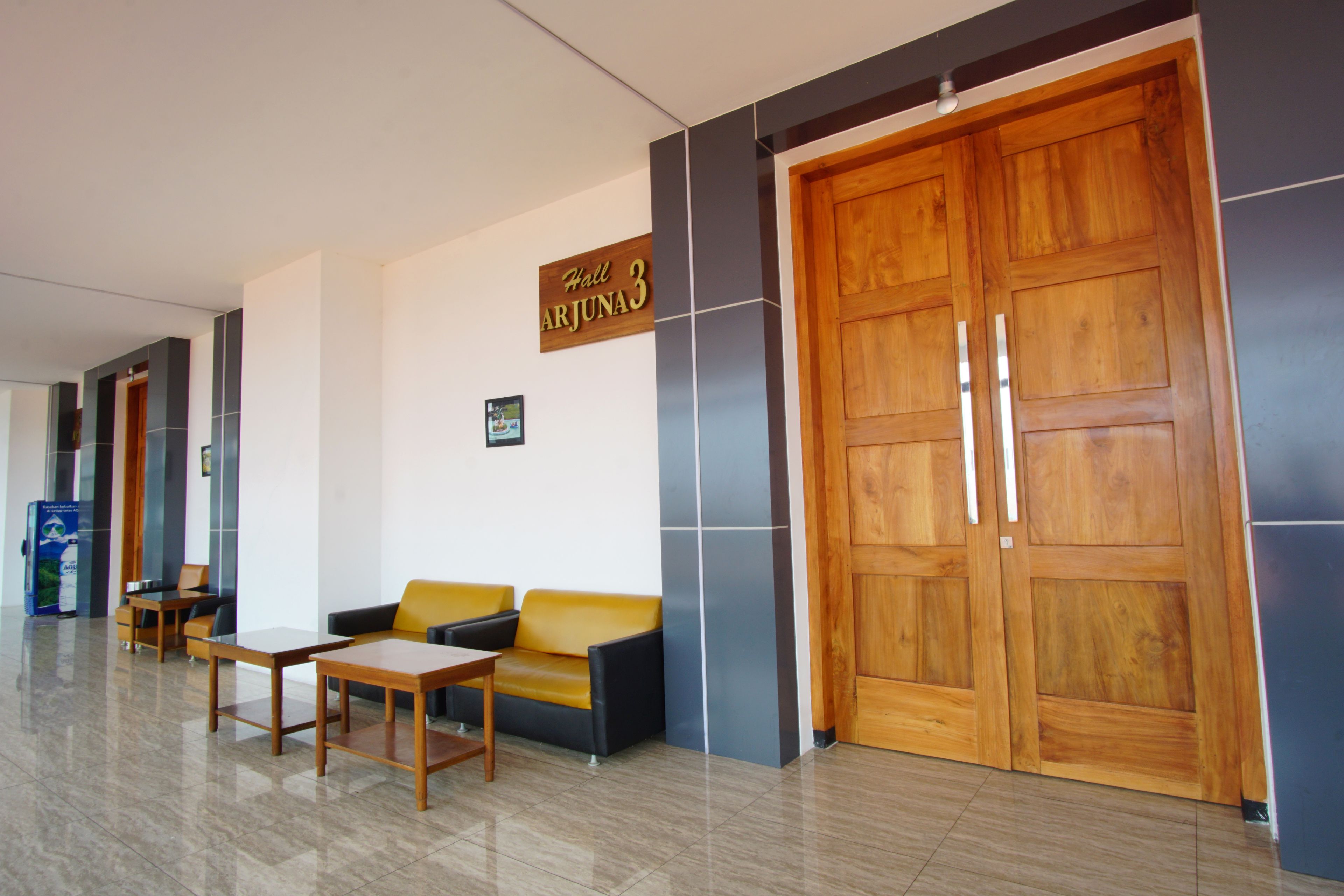 A reception area with a wooden door, a sign reading "Hall ARJUNA 3", a sofa, and a table.