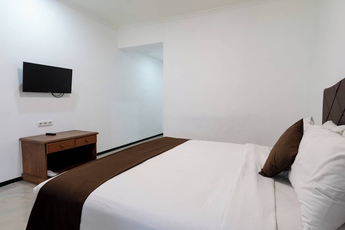 A hotel room with a bed, a wooden nightstand, a television mounted on the wall, and a door leading to another room.