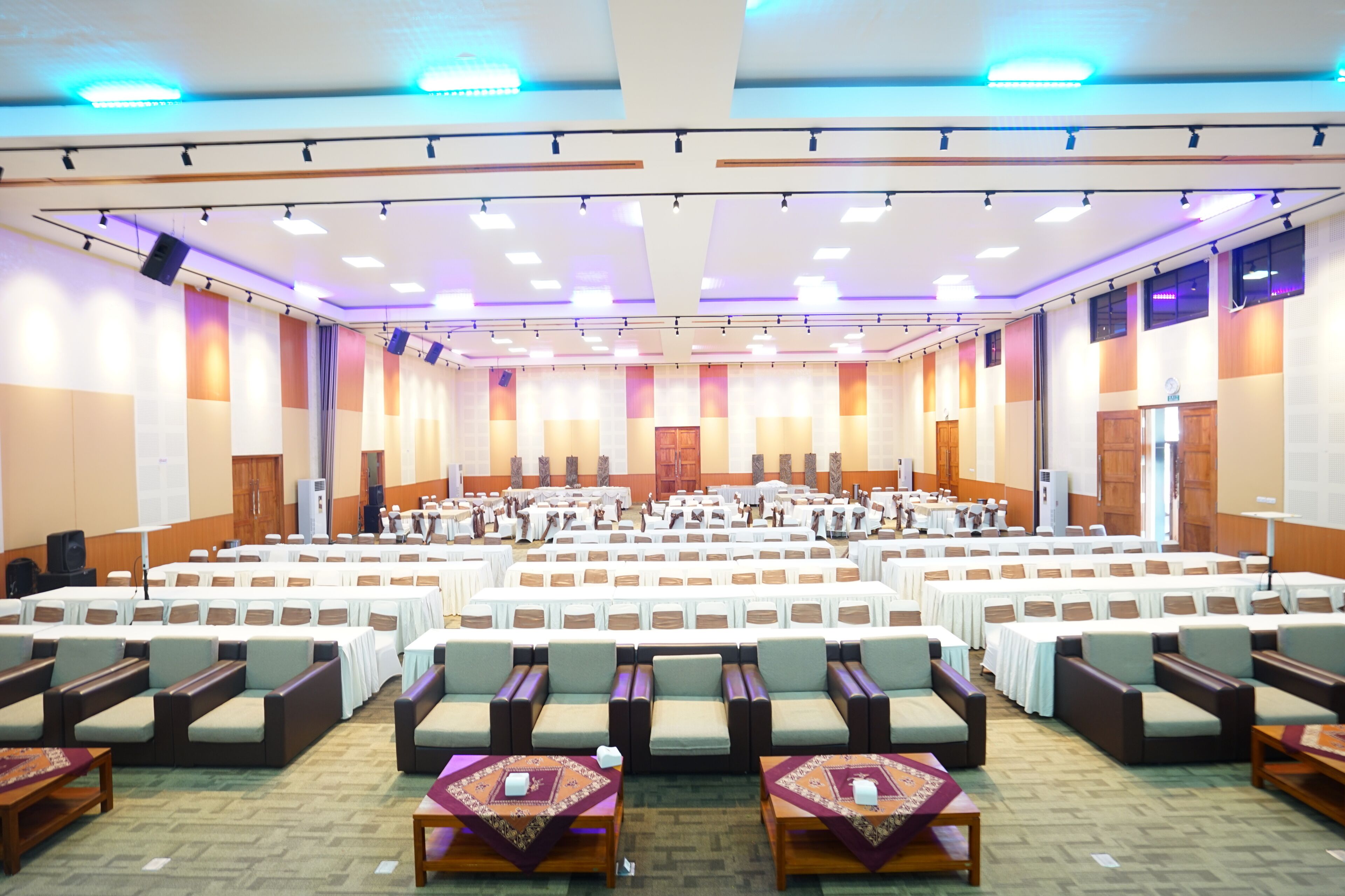A large conference room with rows of tables and chairs, equipped with ceiling lights and a stage at the front.