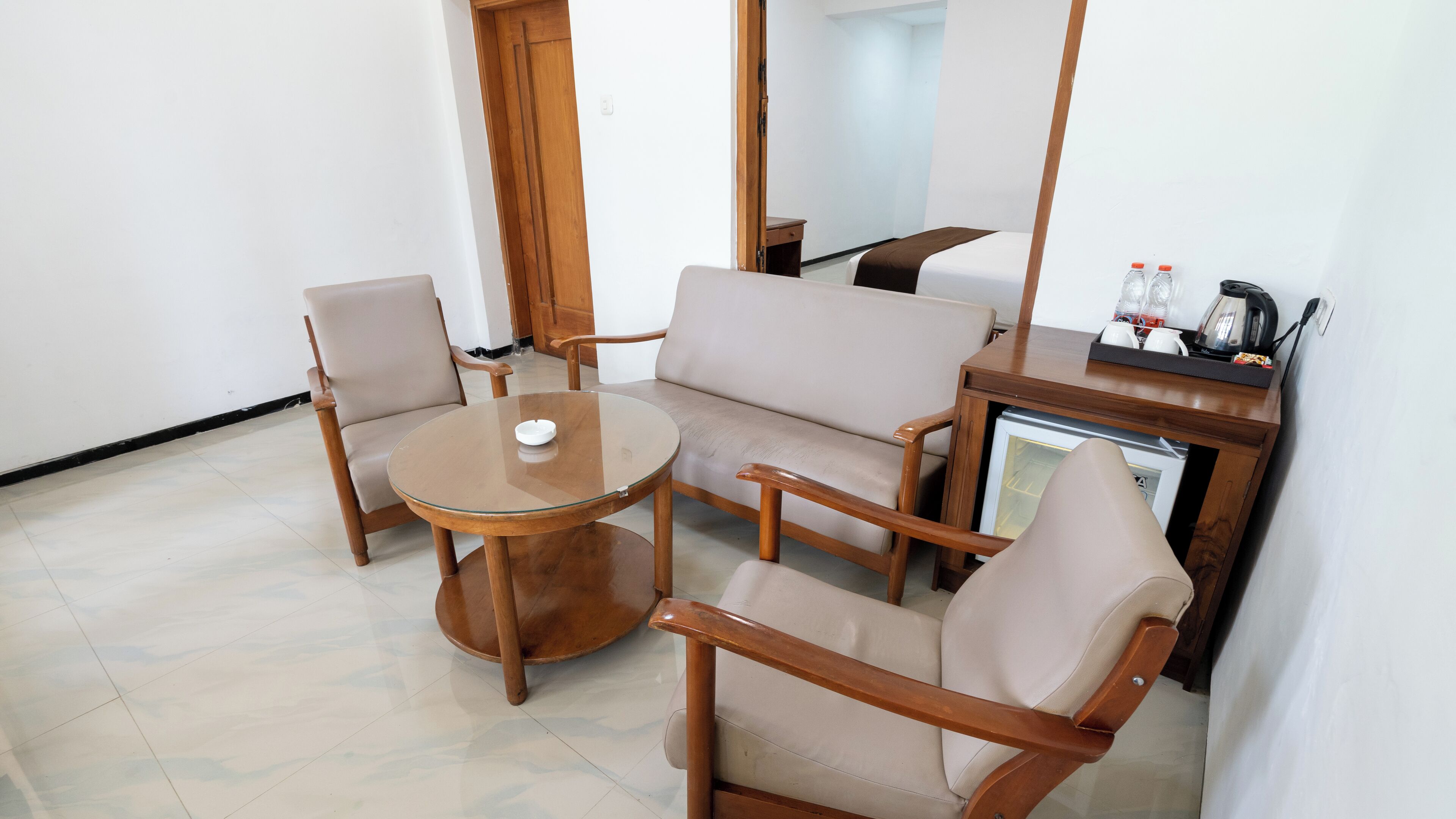 A small, modern living area with a round glass table, two wooden chairs, a sofa, and a small wooden cabinet with a kettle and bottles.
