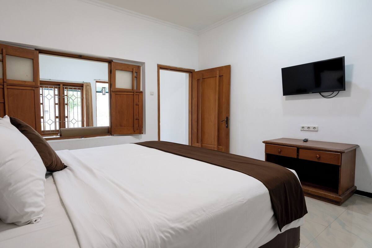 A bedroom with a bed, a wooden wardrobe, a television mounted on the wall, and a window with wooden shutters.