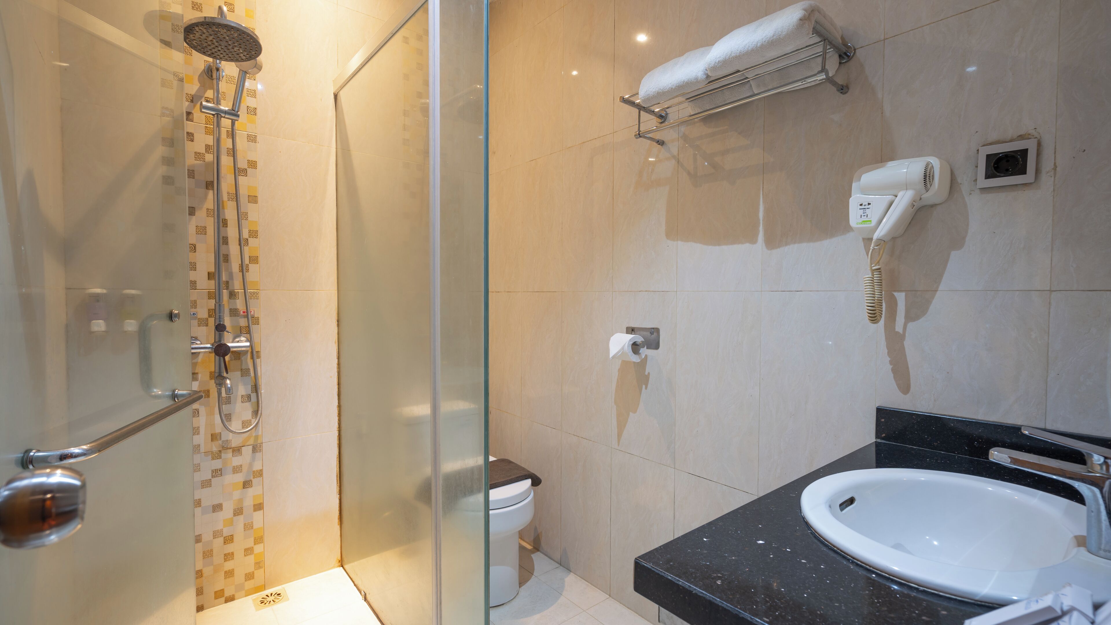 Une salle de bain moderne avec une douche à parois de verre, des toilettes, un lavabo doté d’un plan de travail noir, un porte-serviettes et un sèche-cheveux.
